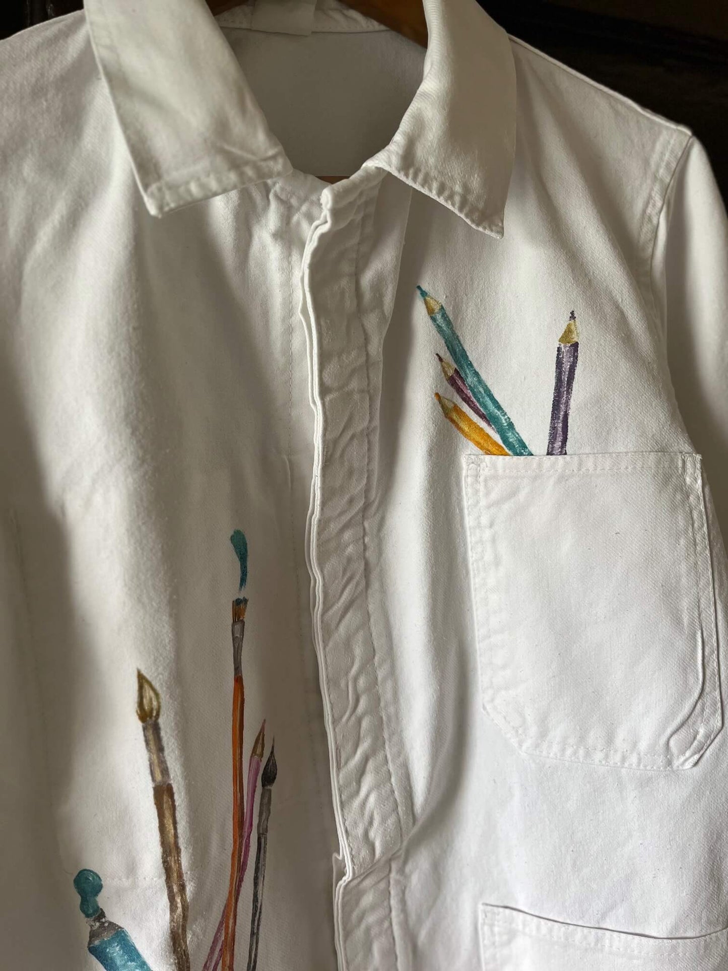 Détails de la veste d’artiste blanche customisée avec des crayons et pinceaux peints à la main sur les poches